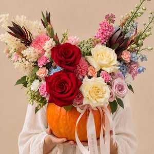 A person in a white long-sleeve top holds a large orange pumpkin filled with a colorful floral arrangement. The bouquet includes red and cream roses, pink and lavender carnations, blue flowers, green foliage, and pale pink ribbons tied around the pumpkin, against a plain beige background.