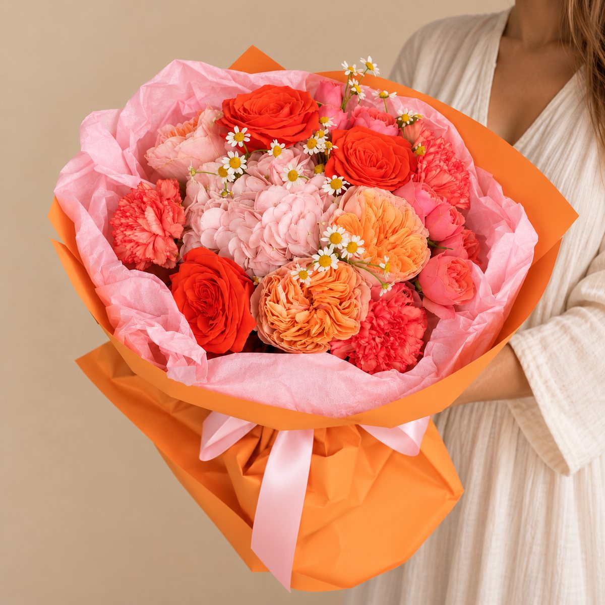 A vibrant bouquet of red, pink, and orange flowers wrapped in pink tissue paper and bright orange paper, tied with a pink ribbon, held by a person wearing a cream-colored robe against a solid beige background.