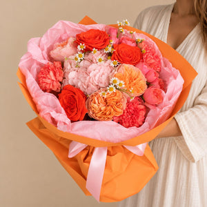 A vibrant bouquet of red, pink, and orange flowers wrapped in pink tissue paper and bright orange paper, tied with a pink ribbon, held by a person wearing a cream-colored robe against a solid beige background.
